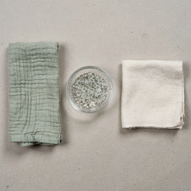 An overhead flat lay of three materials spaced evenly on a grey surface: folded sage-green woven fabric, a small bowl of clear glass beads, and folded off-white cotton fabric.