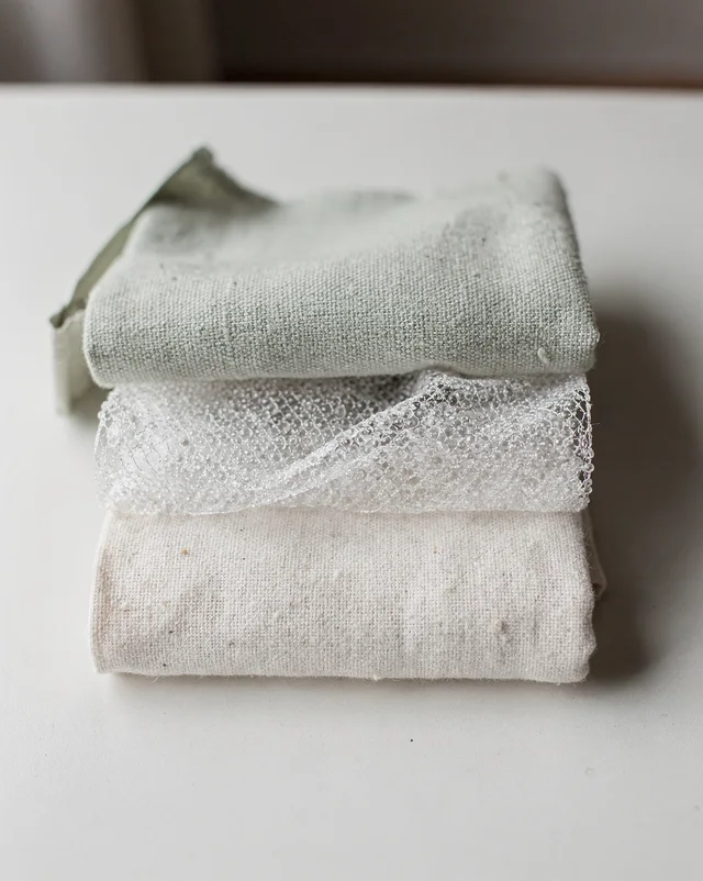 A close-up stack of three fabric layers with visible gaps between them: a pale sage-green woven top layer, a sheer mesh middle layer, and a plain off-white cotton base layer.