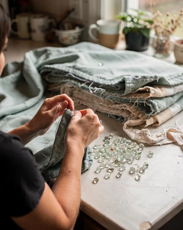 The founder's hands and forearms, unidentifiable, sew a seam at a kitchen table scattered with loose glass beads, mugs and a plant visible in the background.