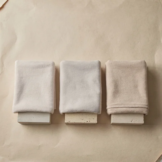 A flat poster-style layout on plain paper: three folded blankets in different shades, each resting on its own small pedestal block, arranged in a row with generous empty space above.