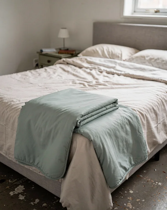 A neatly made bed by a window in daylight, with a folded sage-green quilted blanket resting on top of the cream bedding.