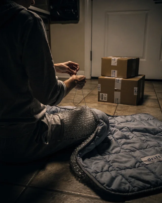 A dim nighttime photo of the founder, unidentifiable and seen from behind, kneeling on a kitchen floor and tying a knot in a bead-filled panel, with two taped ‘drift’ shipping boxes stacked by the door.