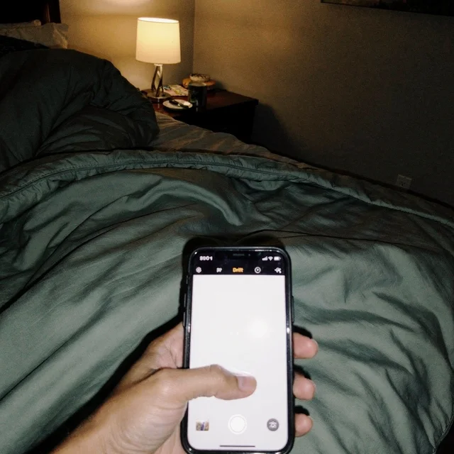 A selfie-style shot of a hand holding up a phone with its camera app open, under a dark green blanket in a dim bedroom with a lit lamp behind. The person's face is out of frame.